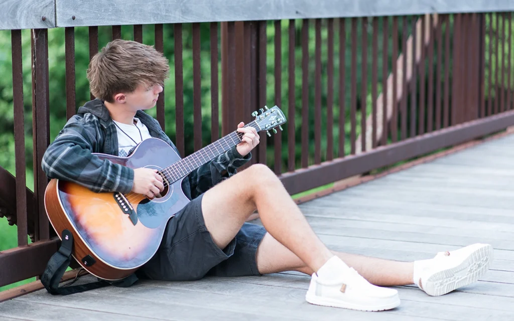 High school senior guy walks away on wooden bridge carrying acoustic guitar — Twin Cities senior portrait photographer