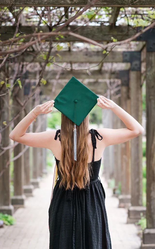 High school senior girl holds green graduation cap under garden arbor — Twin Cities senior portrait photographer