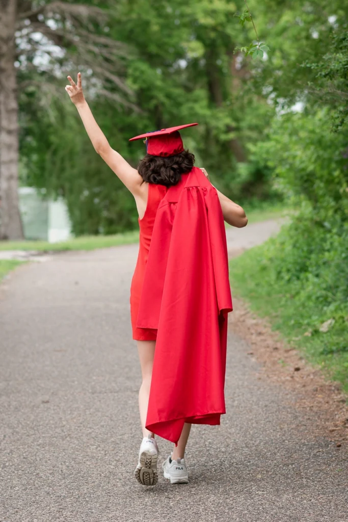High school senior girl walks away in red cap and gown throwing a peace sign — Twin Cities senior portrait photography