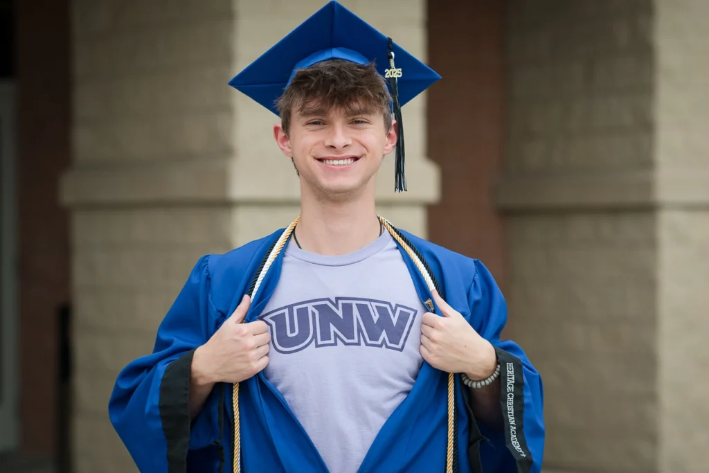 High school senior boy in cap and gown reveals college shirt in fun graduation portrait — Twin Cities senior photography