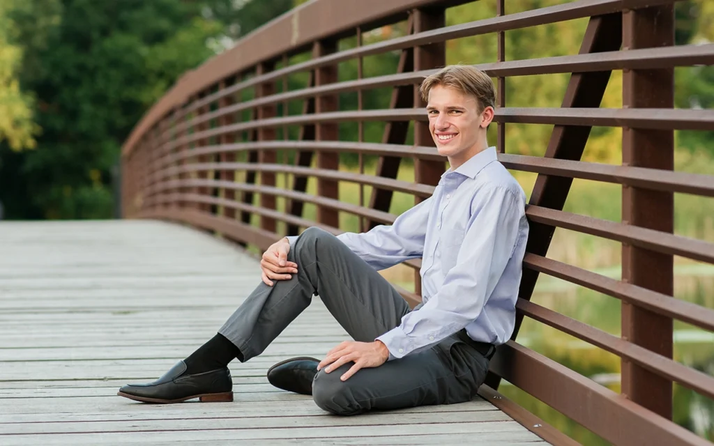 High school senior guy sits relaxed on curved wooden bridge in dress shirt and slacks — Twin Cities senior portrait photographer