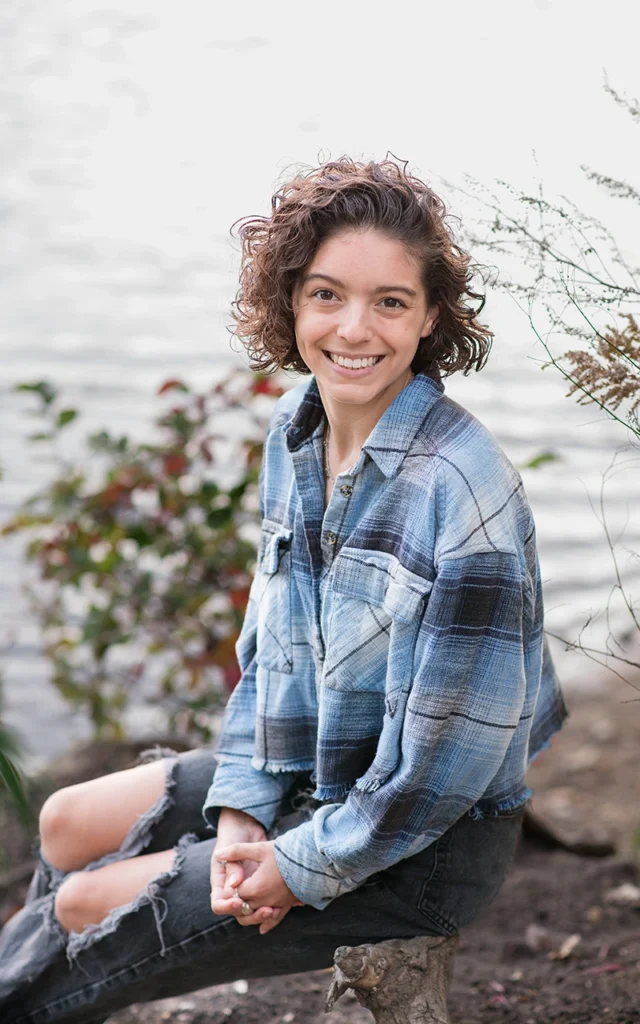High school senior girl sits on log by the water smiling in casual flannel — Minneapolis Minnesota senior portrait photographer