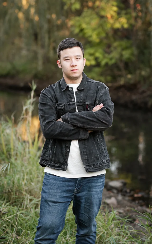 High school senior guy stands arms crossed by autumn creek in black denim jacket — Minnetonka Minnesota senior portrait photographer