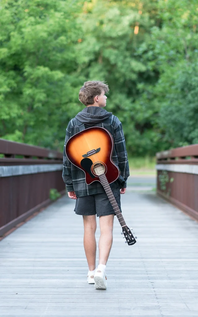 High school senior guy walks away on wooden bridge carrying acoustic guitar — Twin Cities senior portrait photographer