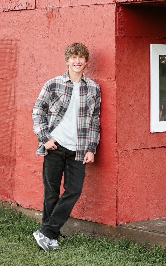 High school senior guy leans against red barn in plaid flannel shirt — Maple Grove Minnesota senior portrait photographer