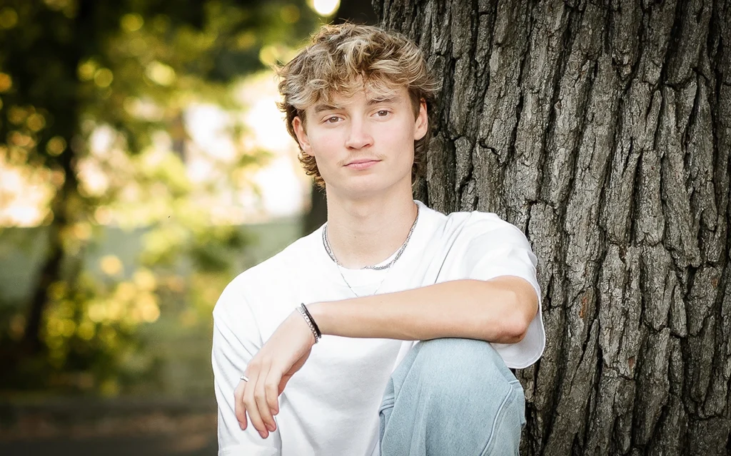 High school senior guy leans against large tree trunk in white tee with golden bokeh background — Champlin Minnesota senior photographer