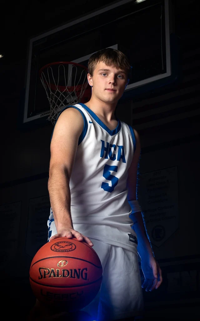 HCA high school basketball player stands with Spalding ball under the hoop in dramatic sports portrait with blue studio lighting
